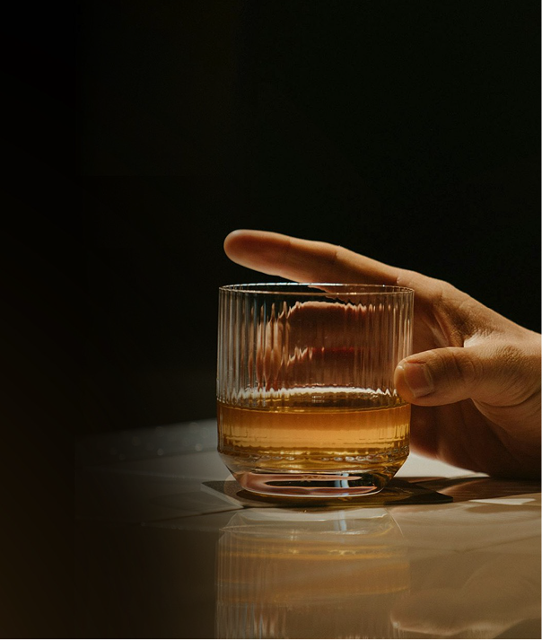 A hand holding a glass of amber-colored liquor on a reflective surface with a dark background, showcasing the elegance and sophistication that Content Capes brings to every moment.