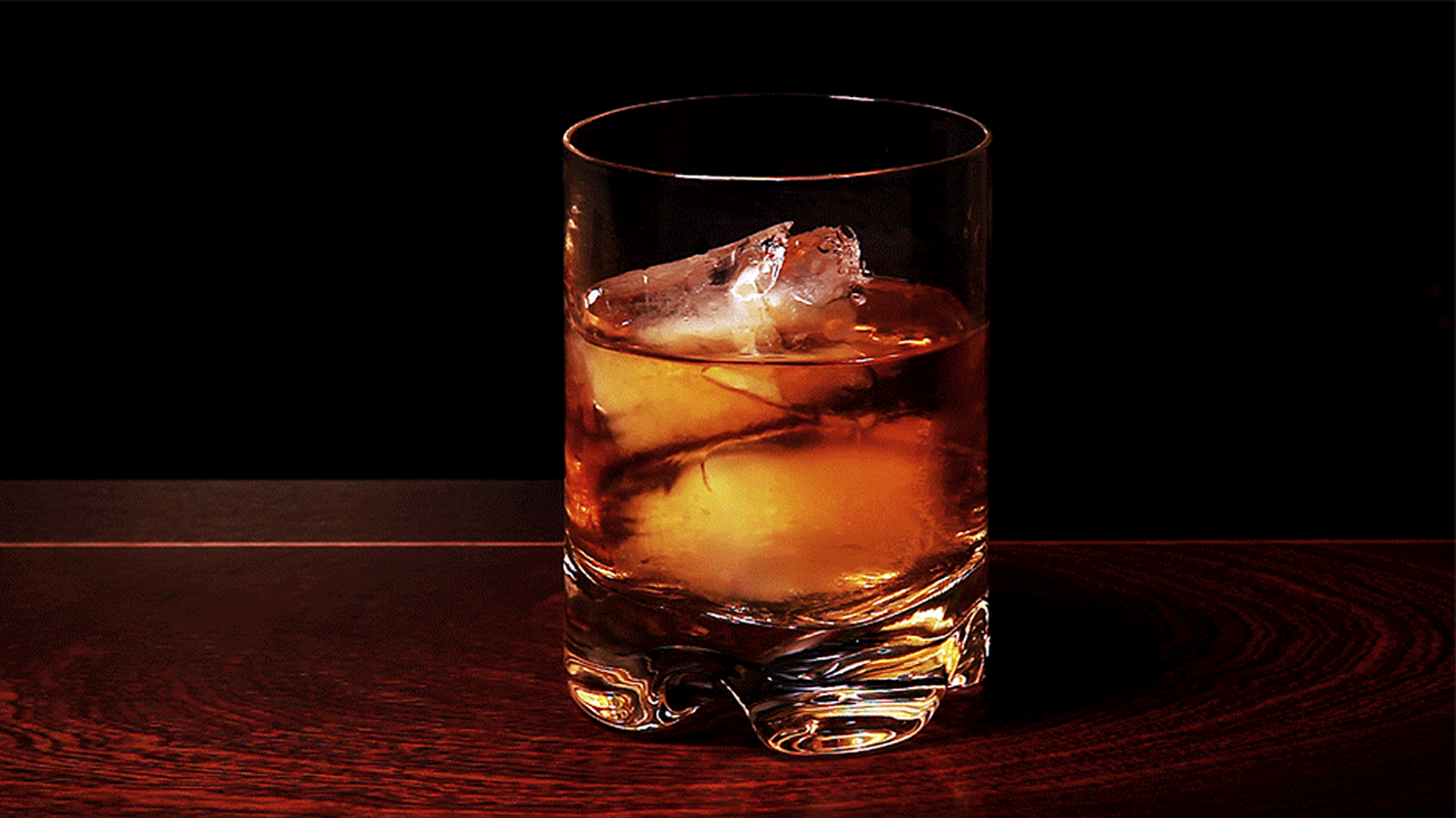 A glass of Meadow Lands whiskey with ice cubes sits on a dark wooden surface, set against a black background, reflecting the brand's dedication to quality distilling.