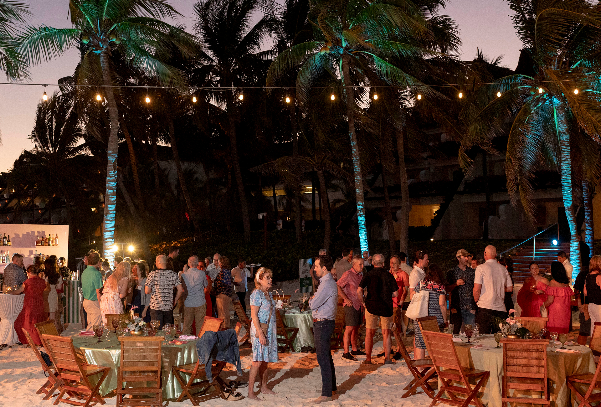 A group of people gather for an outdoor 24 Seven event on a sandy beach at dusk, surrounded by palm trees, string lights, tables, and wooden chairs—celebrating excellence together.