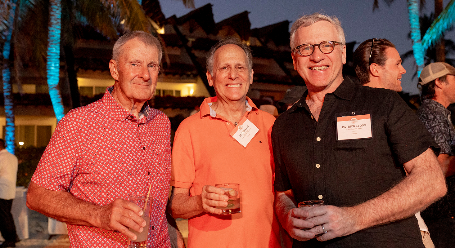 Three men stand outdoors at an evening 24 Seven event, each holding a drink and wearing name tags, with palm trees and building lights in the background, capturing excellence.