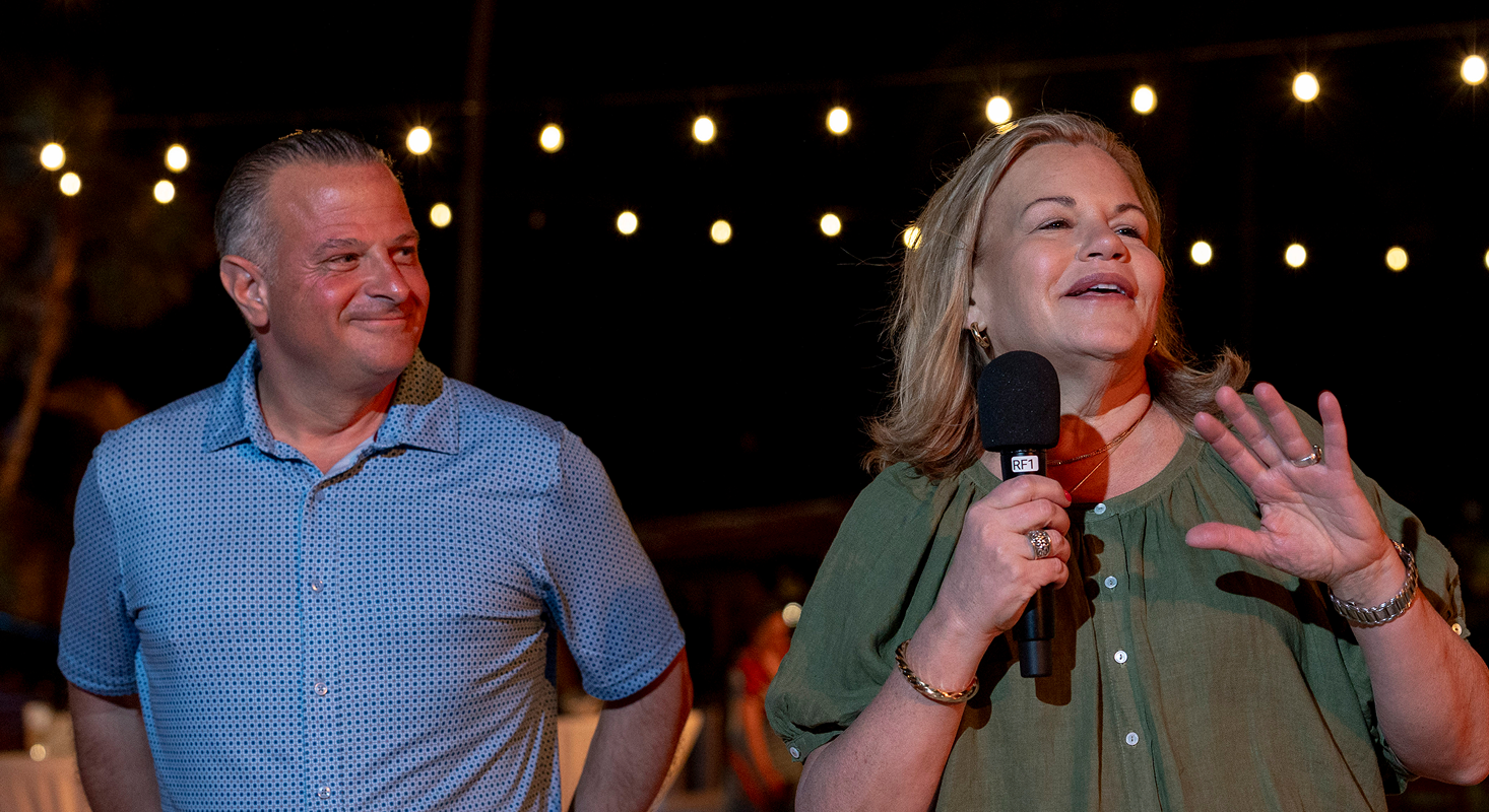 Two adults stand under string lights at night; the woman on the right, celebrating excellence, speaks into a microphone while the man on the left looks at her and smiles.