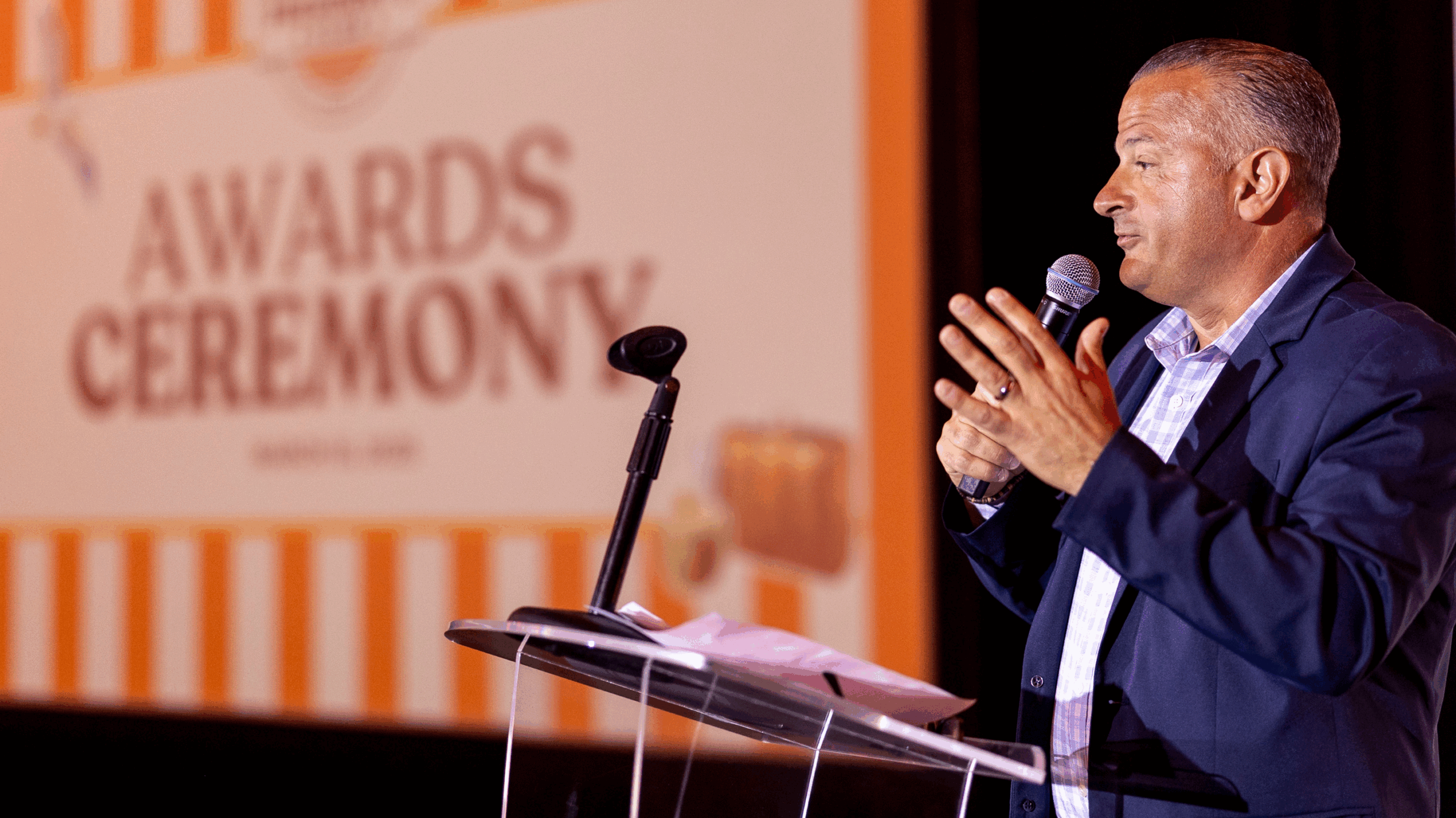 A man in a suit speaks into a microphone at a podium during the 24 Seven President's Club awards ceremony, with a large event sign in the background, celebrating excellence.