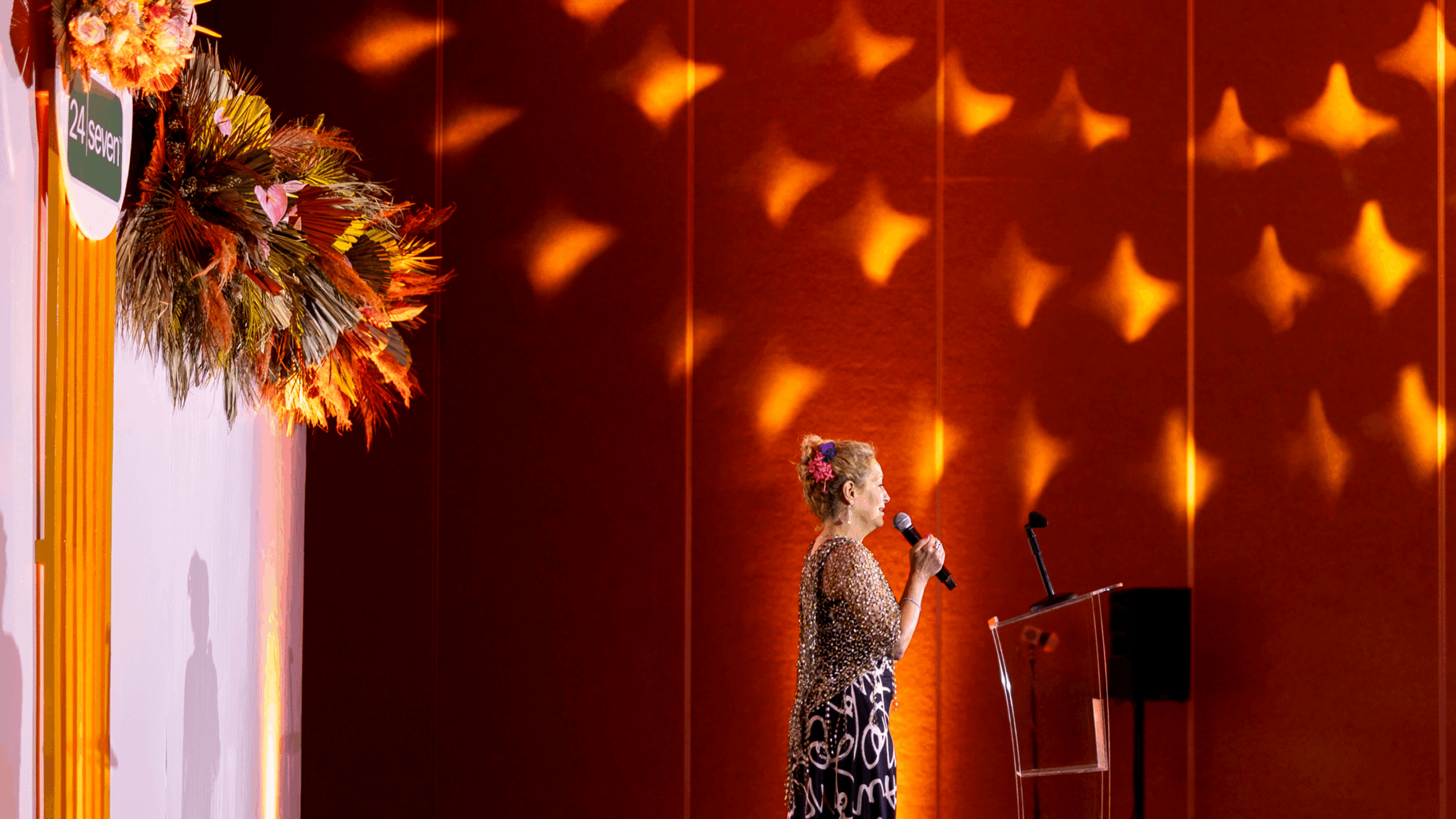 A person stands on stage holding a microphone, speaking near a podium with orange lighting and a decorative floral arrangement on the left, celebrating excellence at the 24 Seven President's Club event.