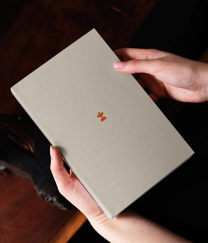 Person holding a closed beige hardcover book with a small, simple orange symbol on the cover.