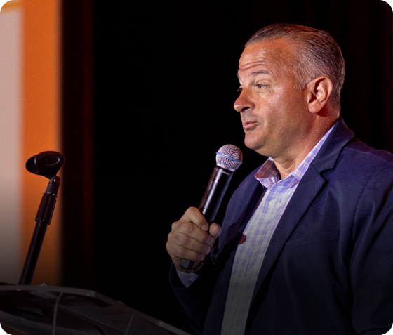 A man in a suit holds a microphone and speaks at a podium against a dark background, capturing excellence at the 24 Seven President's Club event.