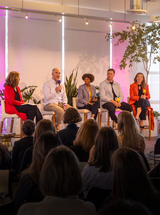 Five people sit on a panel in front of an audience at an indoor event, holding microphones and engaging in discussion. Plants and bright lighting decorate the setting as members of the community of marketers share insights.