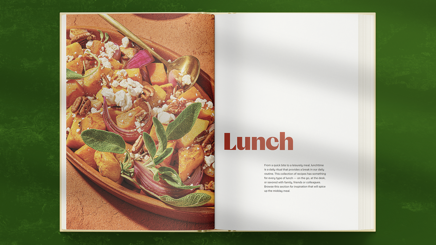 An open cookbook displays a photo of a roasted squash salad with feta, pecans, and sage on the left page and the word “Lunch” with a short description on the right page.