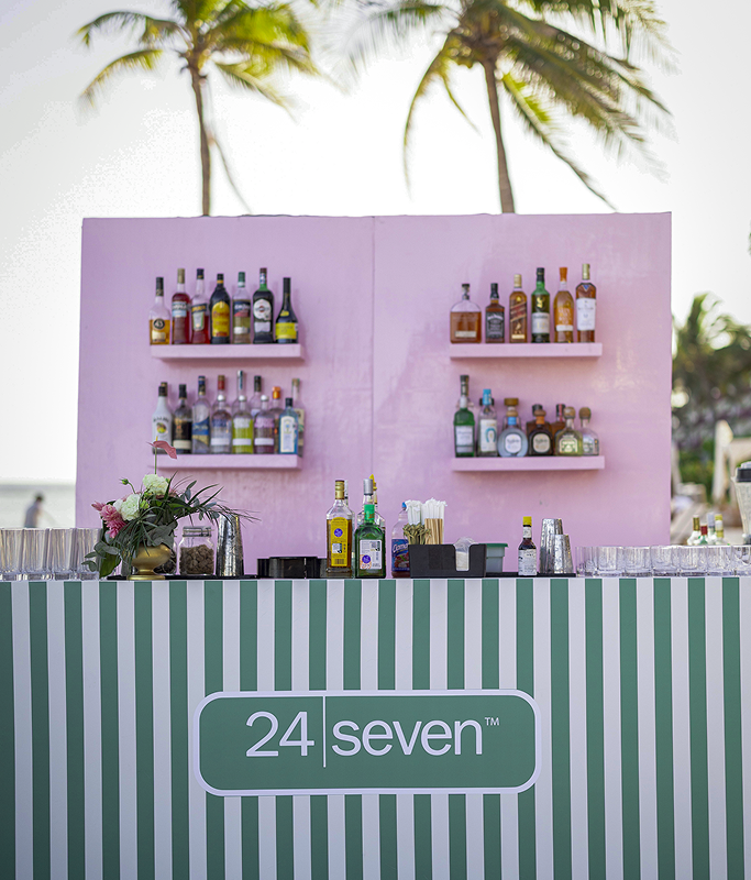 A striped bar counter labeled "24 Seven" stands in front of a pink backdrop with shelves displaying various liquor bottles, embodying excellence; palm trees are visible in the background.