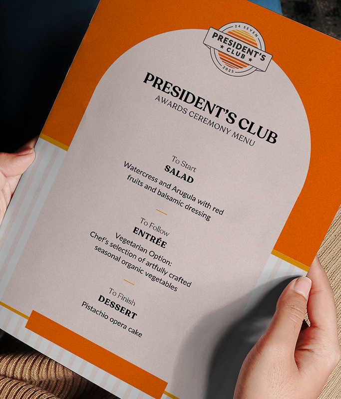 A person holds an orange and white "President's Club Awards Ceremony Menu" from 24 Seven, featuring salad, entrée (and vegetarian option), and pistachio opera cake—a celebration of excellence in every course.