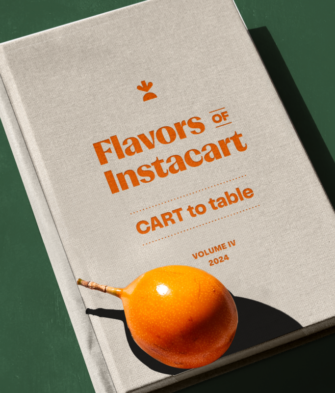 A beige book titled "Flavors of Instacart: CART to table, Volume IV, 2024" rests on a green surface with an orange fruit on its cover, showcasing favorite Instacart recipes from the employee cookbook.