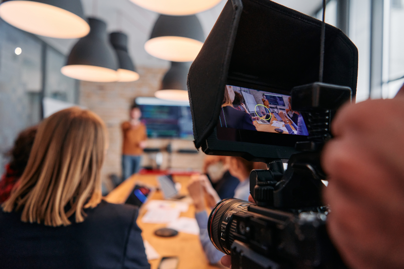 A video camera captures a person giving a presentation to a group seated at a table in a modern office, showcasing effective video storytelling in action.