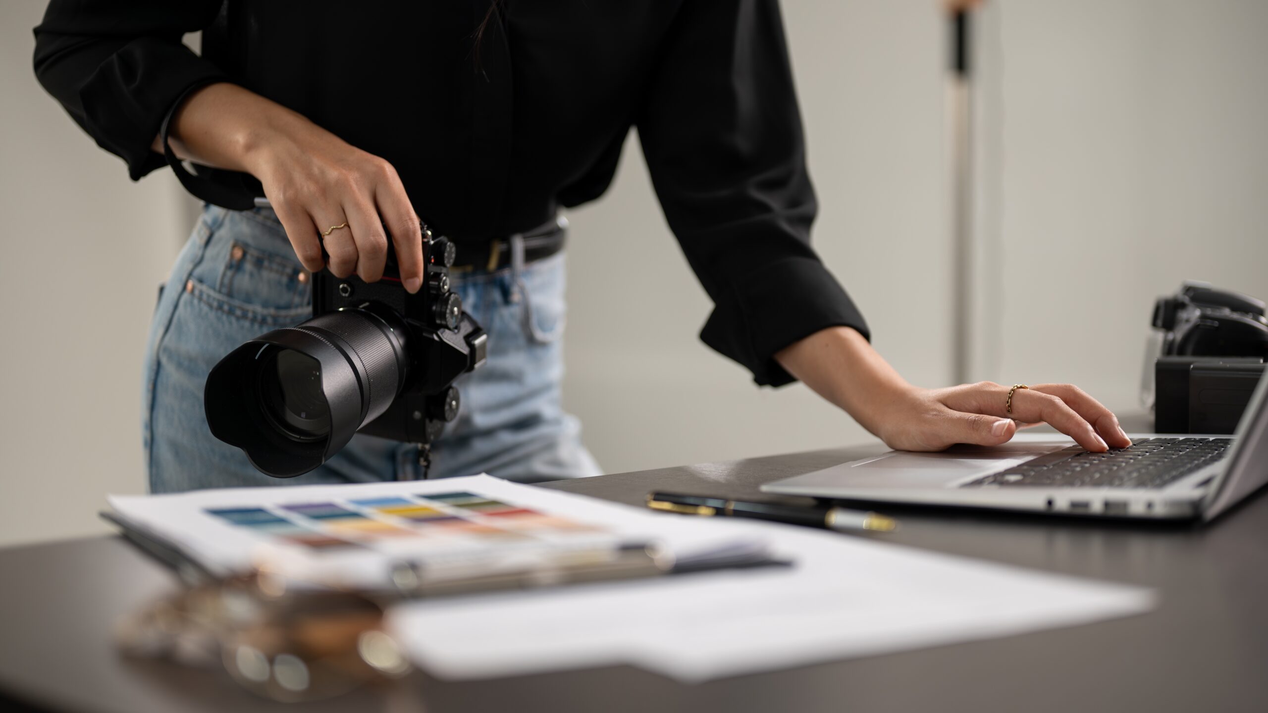 A person holding a camera in one hand and using the trackpad on a laptop with the other, at a desk with papers, a pen, and a color chart—perfectly capturing the authenticity of original photography.