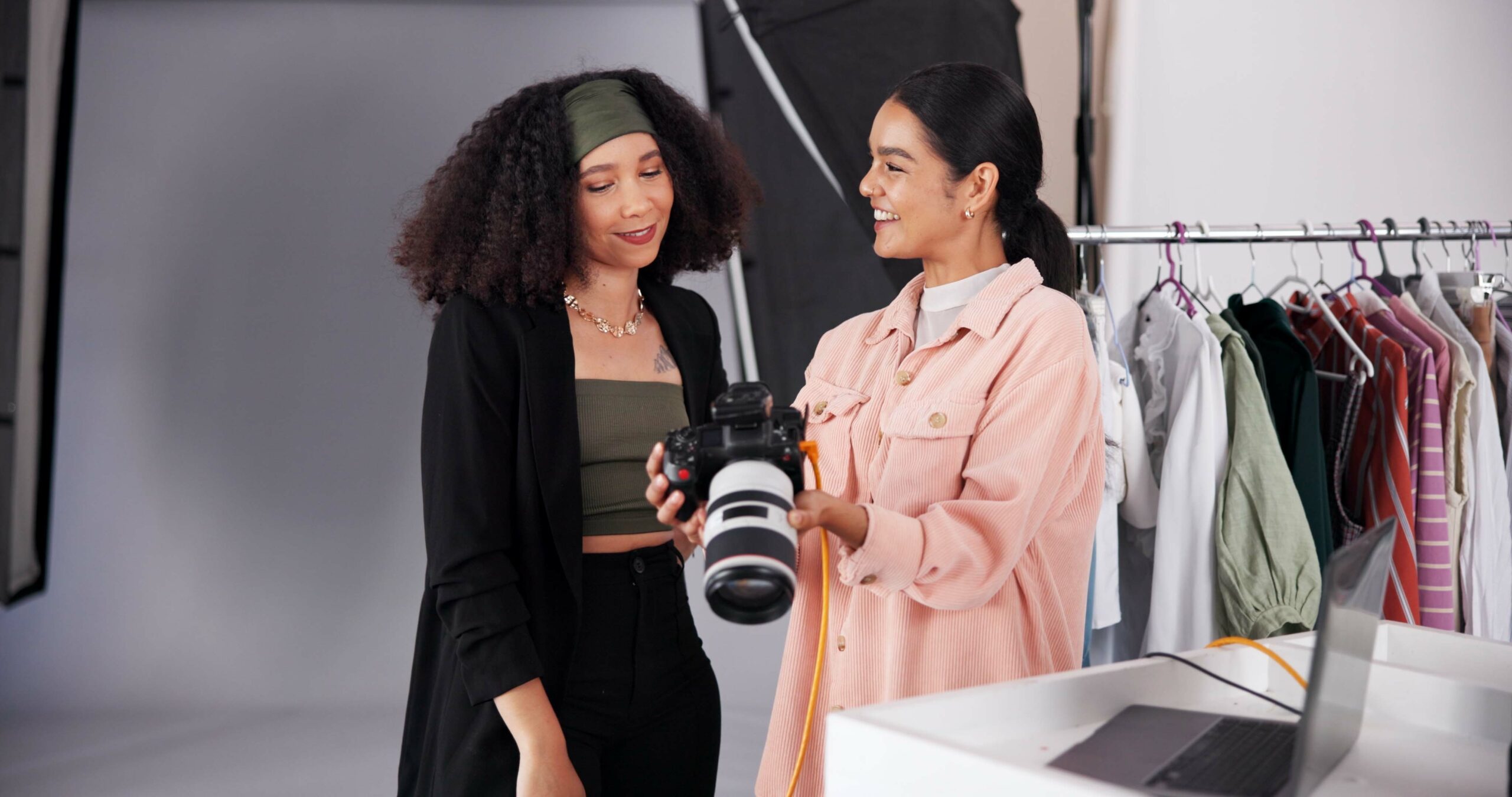 Two women in a studio review images on a digital camera, surrounded by clothing racks and photography equipment, collaborating to enhance brand perception through creative photography.