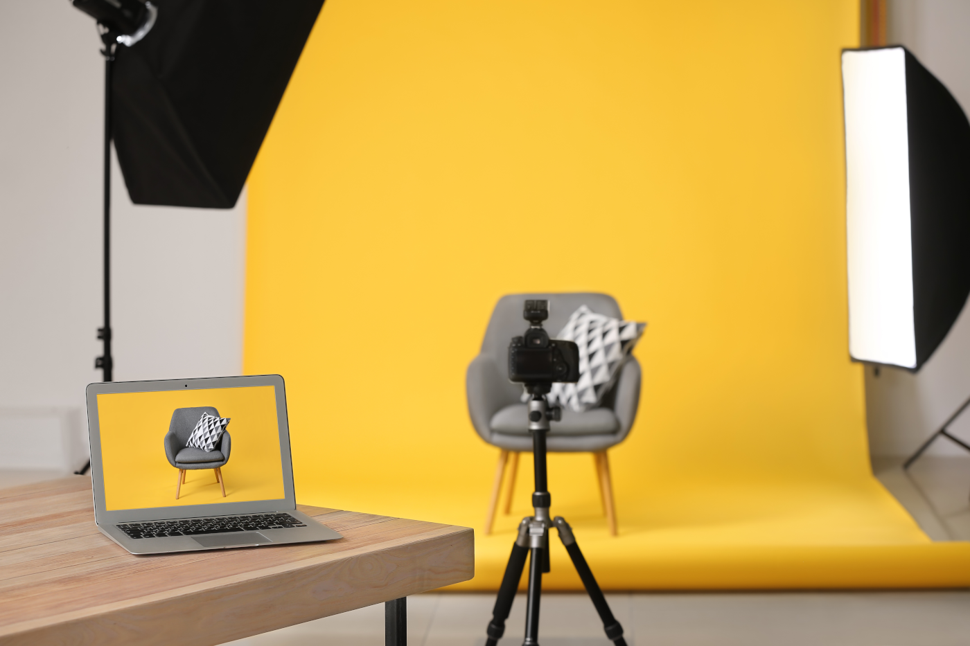 A photo studio setup for a Talking Head Video, featuring a gray chair with a patterned pillow against a yellow backdrop, a camera on a tripod, and a laptop displaying the chair on its screen.
