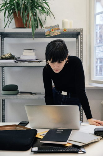 A woman in a black top leans over a desk, looking at her laptop as she works smarter. Notebooks, papers, and office supplies are spread out, with shelves and a plant in the background. Perfect for AI-driven email marketers.