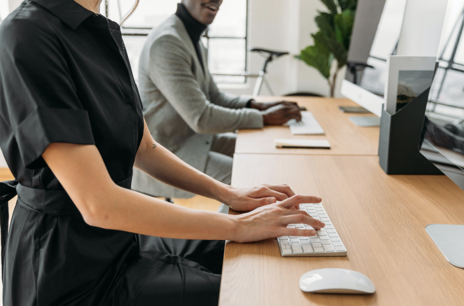 Two people, likely email marketers, sit at a shared desk in a modern office, using desktop computers to work smarter—possibly leveraging AI tools for greater efficiency.