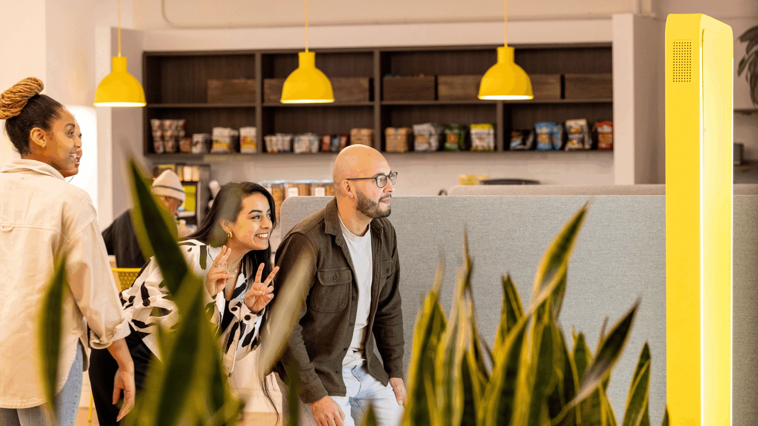 Three people pose and smile in an office space, standing in front of a tall yellow kiosk, showcasing employer brand photography with plants and shelves of snacks in the background.