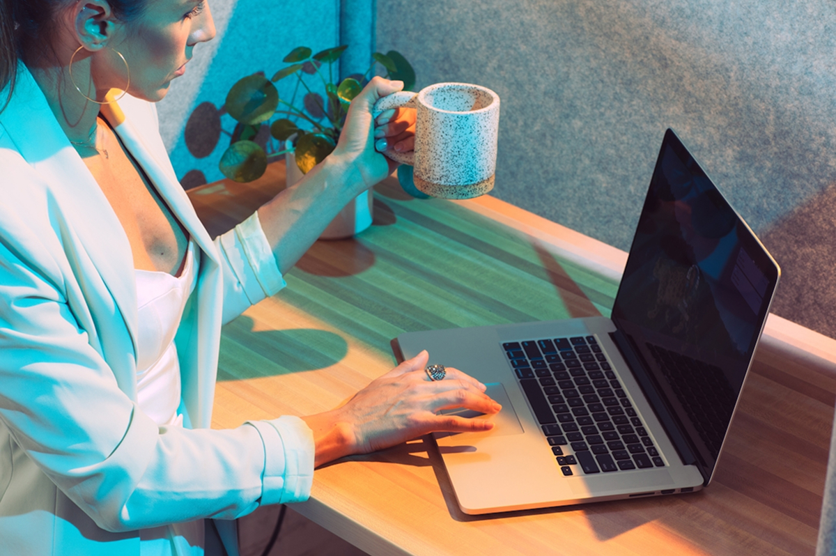A woman sits at a desk using a laptop with one hand while holding a mug in the other, reviewing best practices for web design optimization. A small potted plant is on the desk beside her.