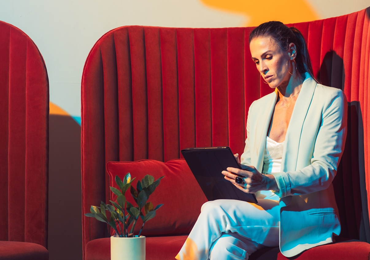 A woman in a white suit sits on a red velvet bench, looking at a tablet, with a potted plant beside her—possibly reviewing Landing Page Design trends for 2025 to boost conversions.