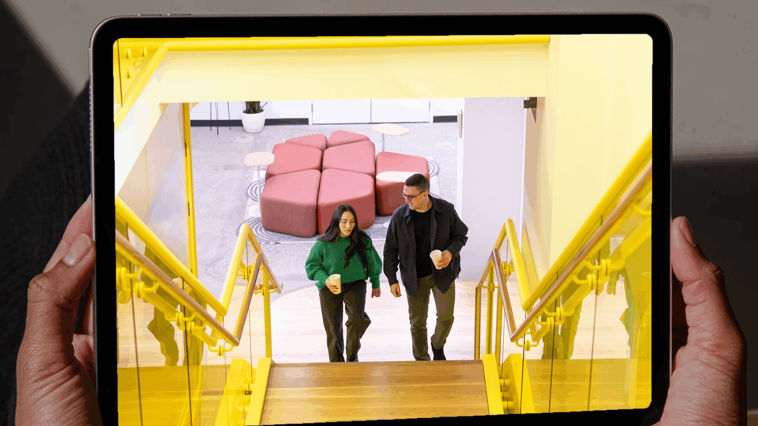 Two people holding coffee cups walk up a yellow staircase, as seen on a tablet screen held by a person—an engaging example of Employer Brand Photography. Pink seating is visible in the background.