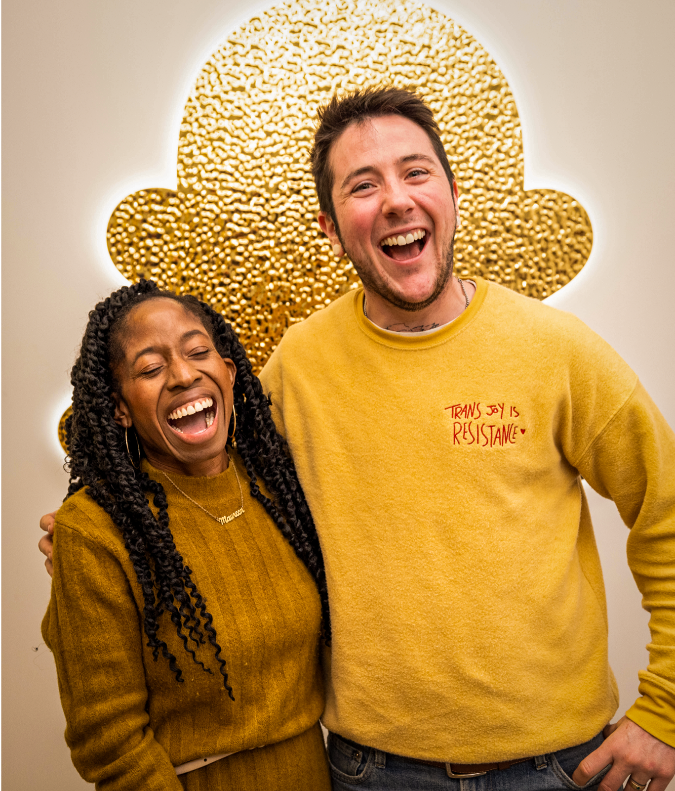Two people stand side by side, smiling and laughing in front of gold, textured wall art. One wears a yellow sweater with "TRANS JOY IS RESISTANCE" text—capturing authentic moments for employer brand photography.