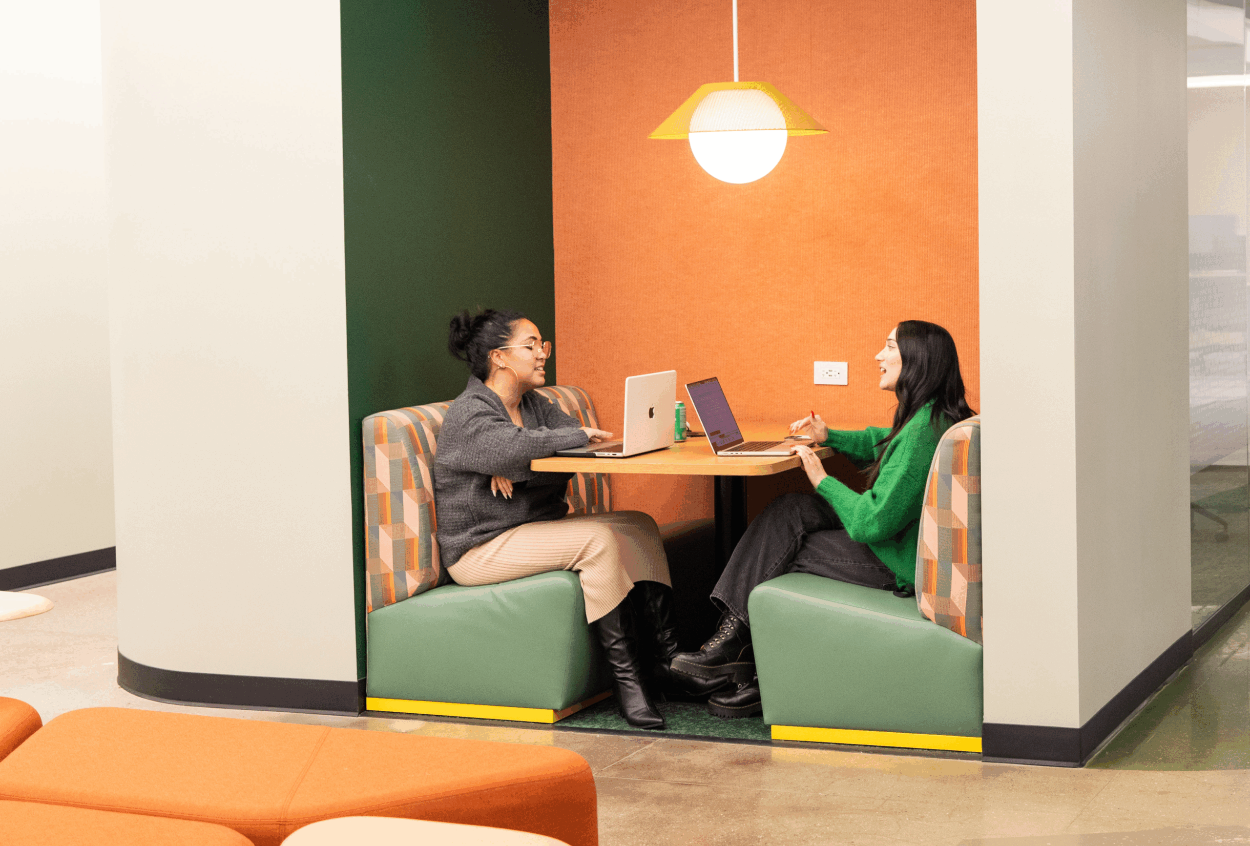 Two women sit across from each other at a table in a booth, working on laptops and discussing ideas in a modern, brightly lit office—capturing authentic employer brand photography.