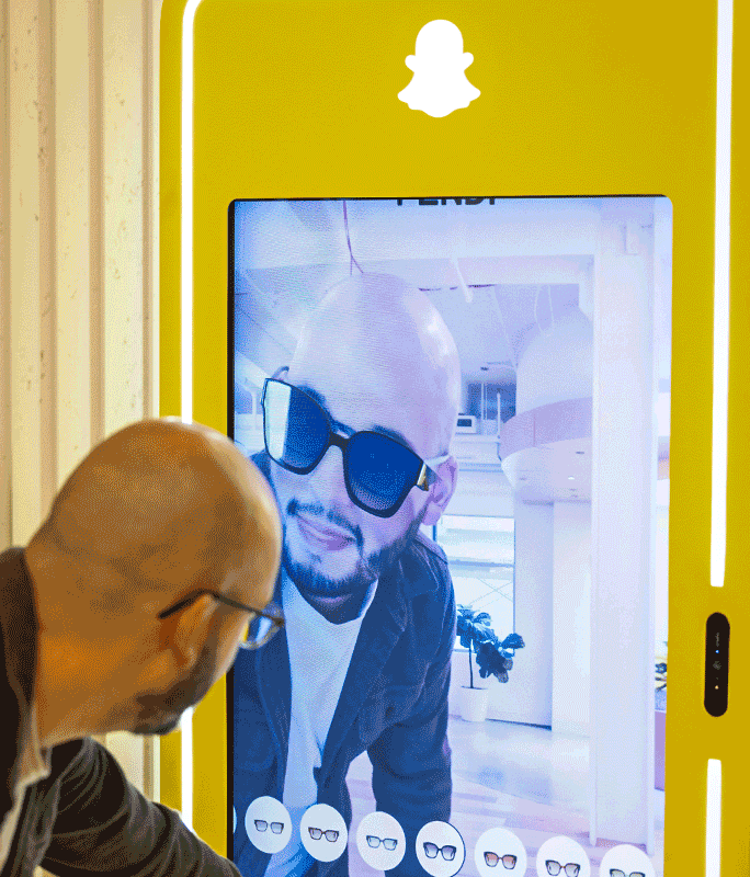 A man uses a Snapchat kiosk to virtually try on sunglasses, looking at his image on a large yellow touchscreen display.