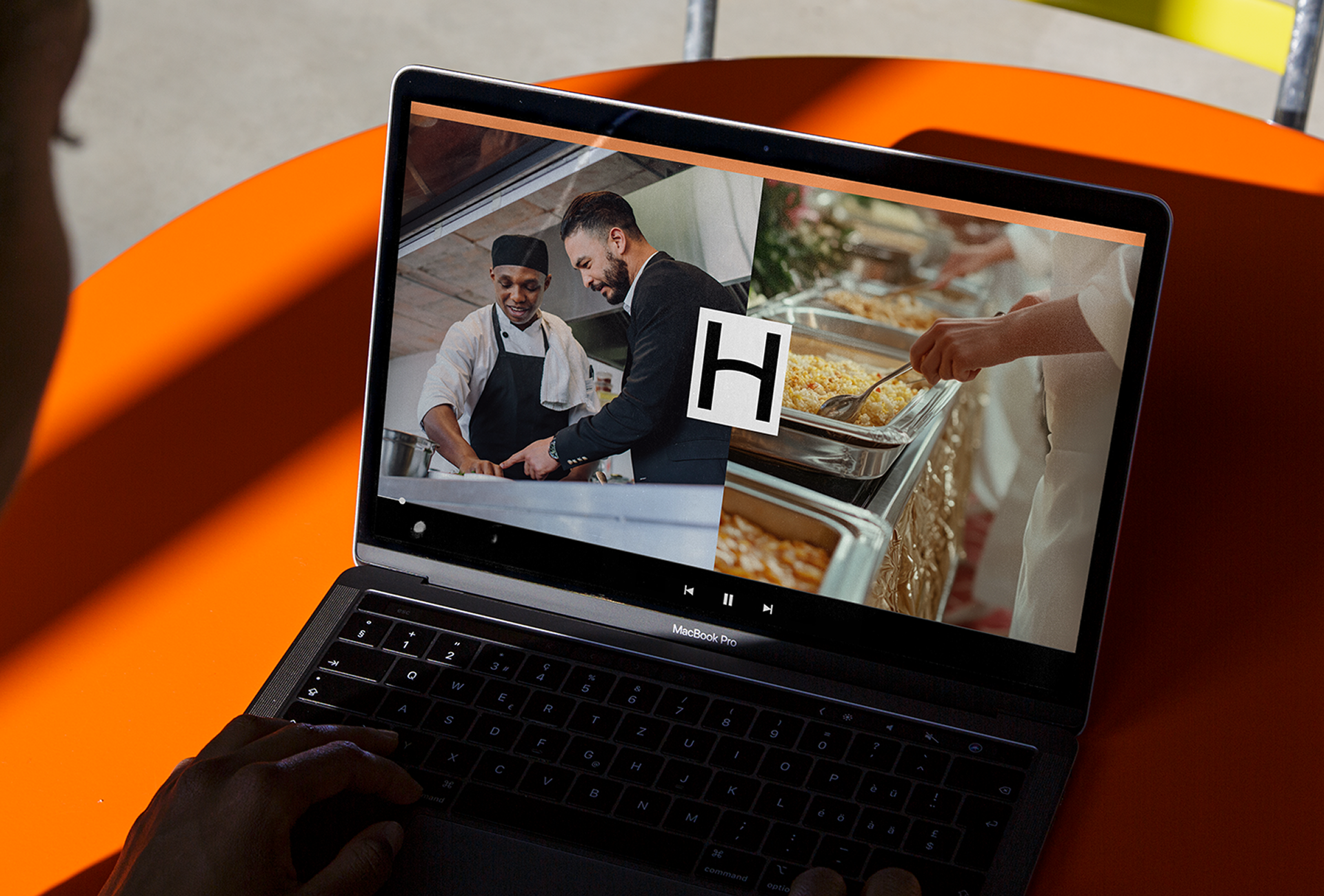 A person watches a video on a laptop featuring chefs preparing food and serving buffet trays, with a large "H" logo in the center—highlighting TAKKT Foodservices brands in action.