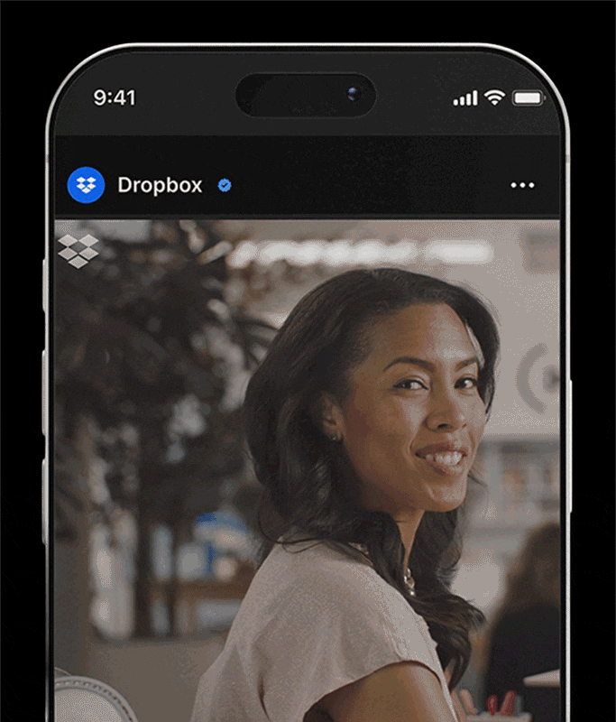 A phone screen displays a Dropbox Dash social media post featuring a woman with long hair smiling and looking over her shoulder amid the creative chaos of an office setting.