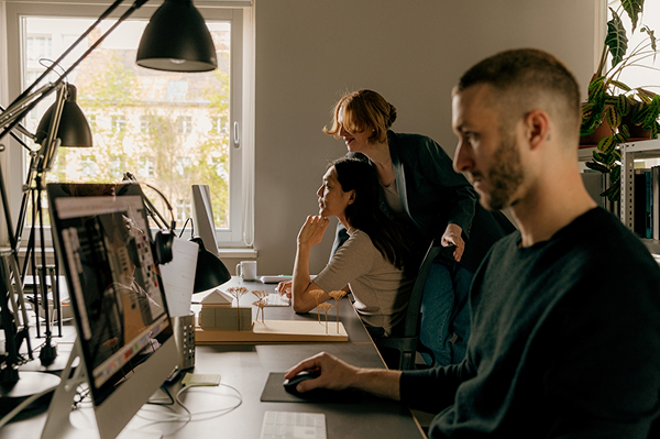 Three people work at desks in a sunlit office. One person uses a computer, while two others collaborate and compare Elementor Pro vs Gutenberg on a screen together in the background.