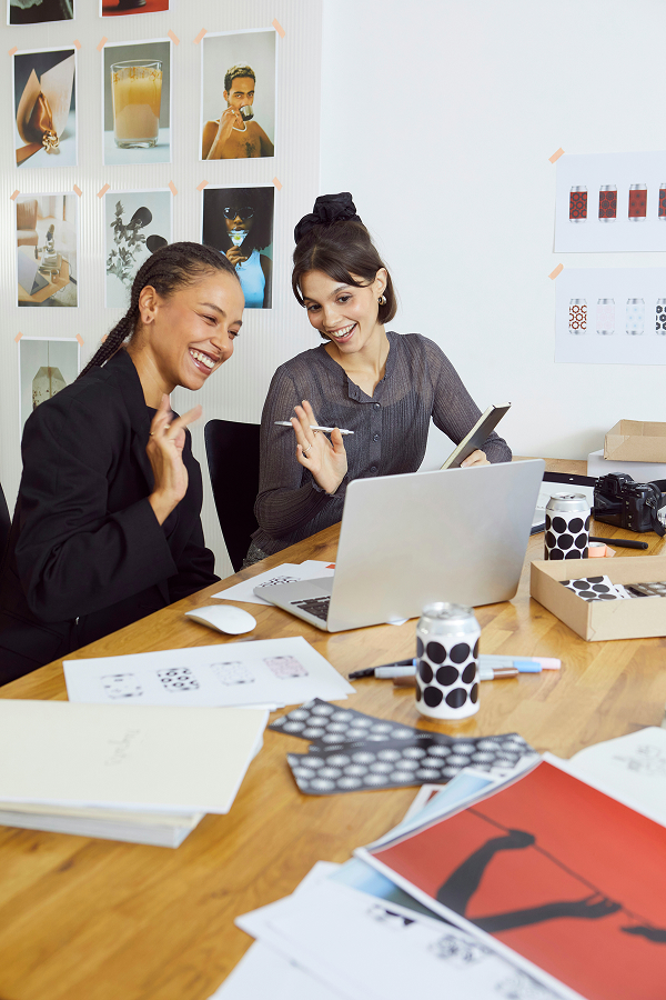 Two women sit at a desk covered with papers and PowerPoint design materials, smiling and waving at a laptop during a video call in a modern office space.