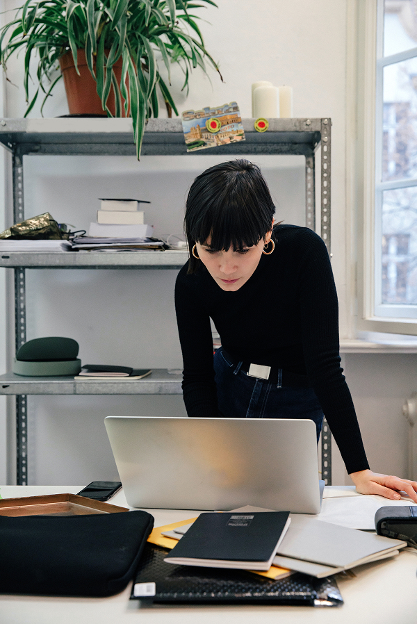 A woman in a black sweater stands at a desk, leaning over a laptop with notebooks, papers, and Pitch Deck materials scattered around, in a room with shelves and a window.