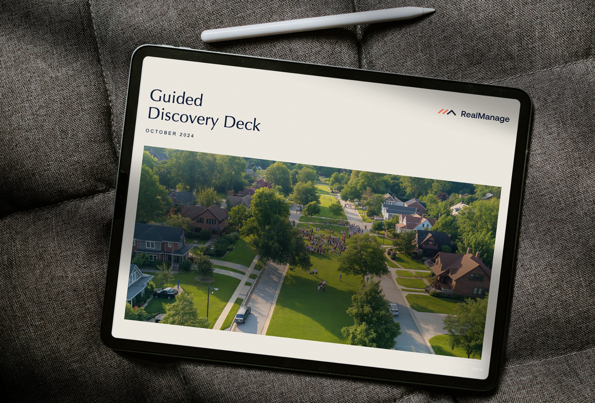 An iPad displaying a presentation titled "Guided Discovery Deck" with an aerial photo of a suburban neighborhood, placed on a gray cushion with a stylus above it, highlights RealManage's brand identity in property management SaaS.