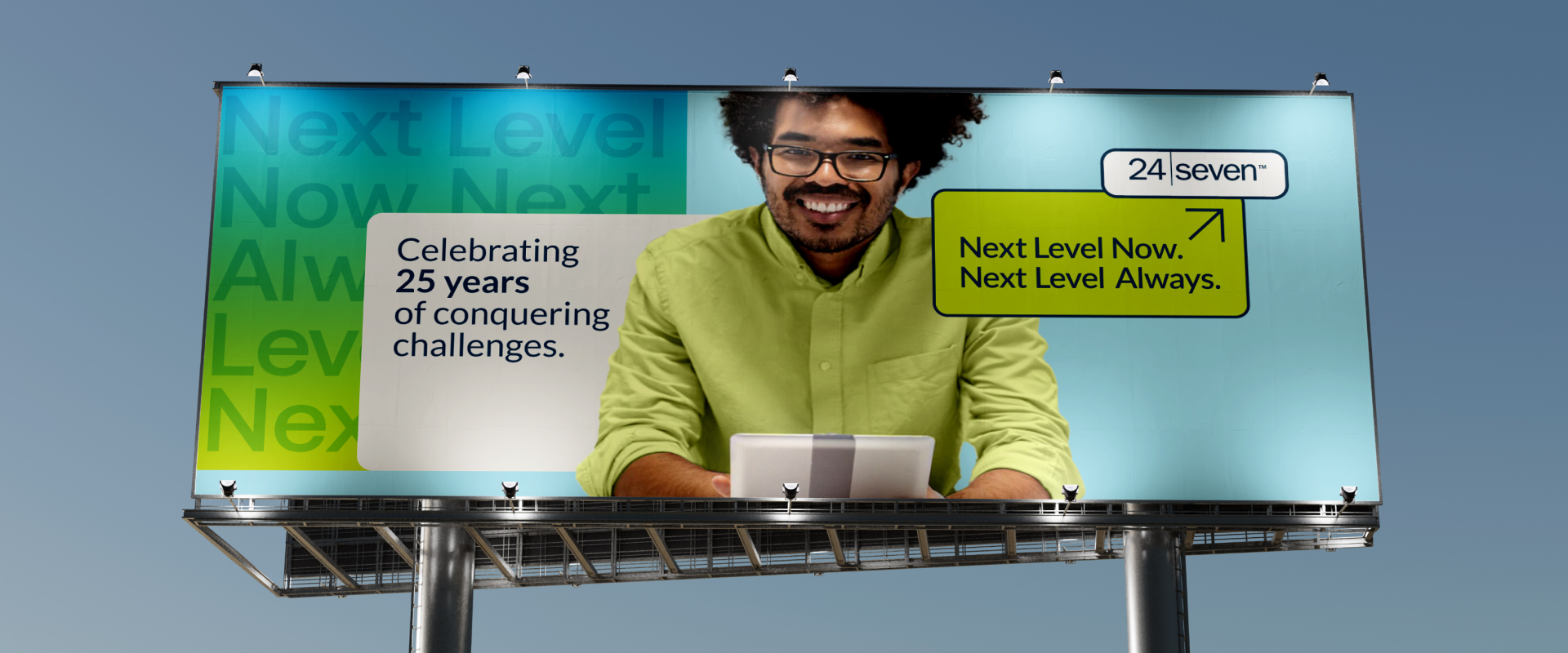 A billboard featuring a smiling person holding a tablet, with text: “Celebrating 25 years of next-level success. Next Level Now. Next Level Always. 24 Seven.”.
