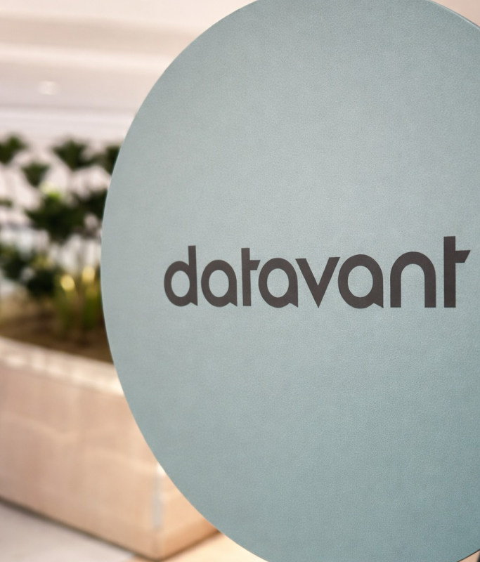 A circular sign with the word "Datavant" in lowercase letters, positioned indoors with blurred plants in the background, reflects a unified approach and hints at executive experience within the space.