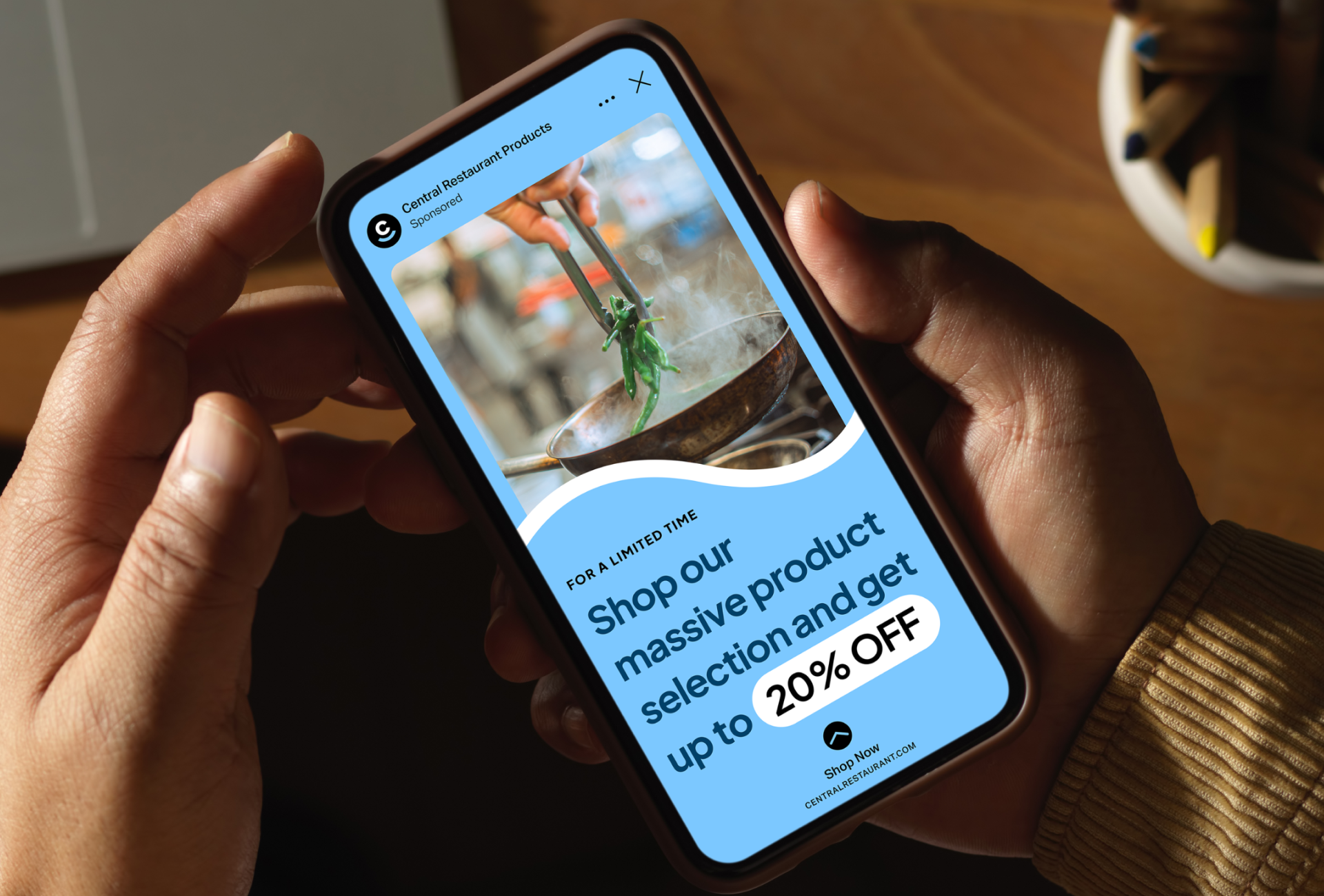 A person holds a phone displaying an ad for Central Restaurant Products, part of the TAKKT Foodservices brand mix, offering up to 20% off on a wide selection, featuring hands cooking with a wok.